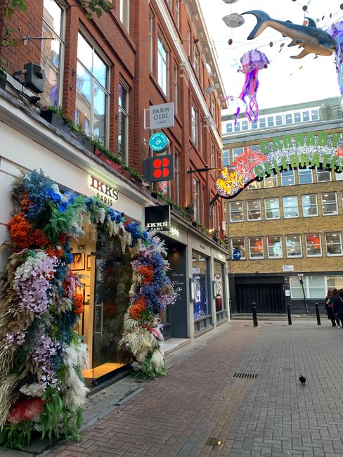 A street scene on Carnaby Street in Soho, showing a row of retail shops including a Starbucks Coffee store on the ground floor of a three-storey building with a purple and red façade, and a Menchaz f