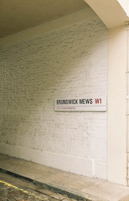A white brick wall with a street sign mounted on it displaying 'BRUNSWICK MEWS W1' in black and red text, indicating a location in Westminster. The wall is part of an interior or exterior urban settin